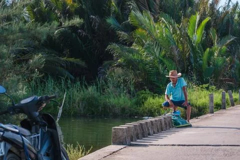 Asian man in a straw hat resting on a concrete bridge over a river Stock-Fotos