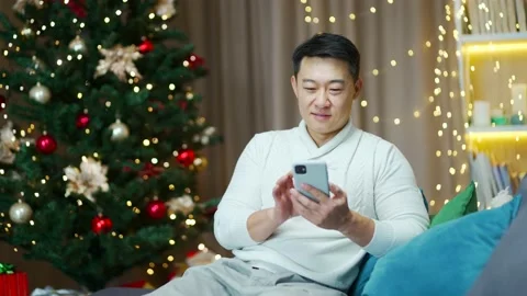 Asian man uses smartphone sitting at home on sofa during winter new year holiday Stock Footage 219912205