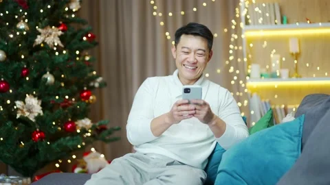 Asian man uses smartphone sitting at home on sofa during winter new year holiday Stock Footage 219912226