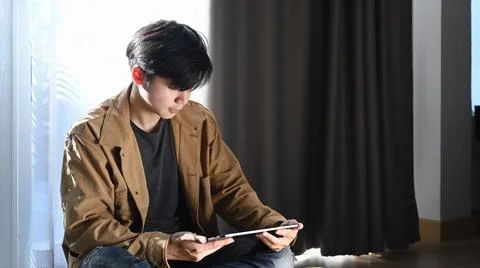 Asian man using digital tablet and sitting on wooden floor at home. Stock Photos