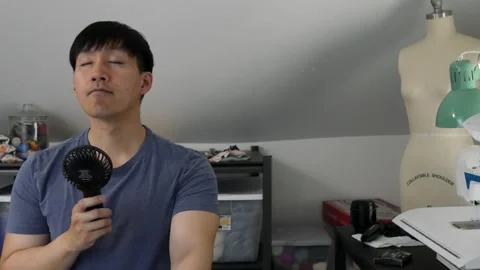 Asian man using a handheld fan on himself in a hot room Stock Footage 247874280