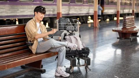 An Asian man is using his digital tablet on a bench while waiting for his t.. Stock Photos