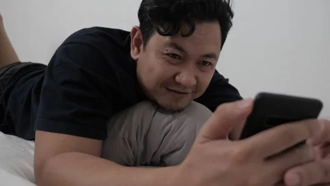 Asian man using phone on bed, reading chat message while lying on matress i.. Stock Footage 296004407