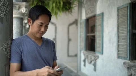 Asian man using smartphone and leaning against Roman-style cement post. Stock Footage 126506184