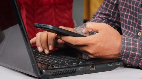 Asian Man using a smartphone while working on a laptop at a desk. Stock Footage 348653223