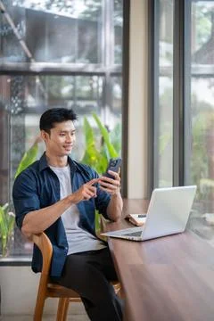 Asian man using smartphone while sitting with laptop at cafe table. Stock Photos