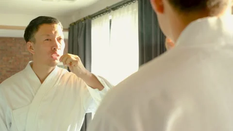 Asian man in white bathrobe brushing teeth standing in front of mirror, oral Stock Footage 232562579