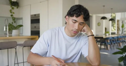 Asian man writing in notebook at home kitchen table, pausing, tapping copper Stock Footage 336175237