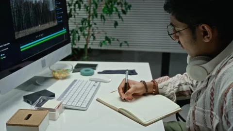 Asian Man Writing in Notebook While Editing Cgi Video on Computer Stock Footage 348350173