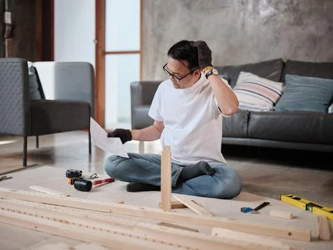 Asian mature man assembles flat-pack furniture with DIY tools and instruction. Stock Photos