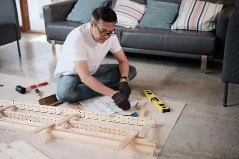 Asian mature man self-assembles flat-pack wooden furniture step by step. Stock Photos