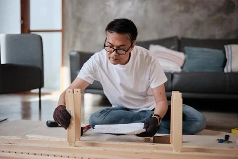 Asian mature man self-assembles flat-pack wooden furniture step by step. Stock Photos
