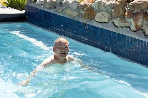Asian middle-aged man smiling, floating chest-up in backyard pool by blue mosaic Foto stock