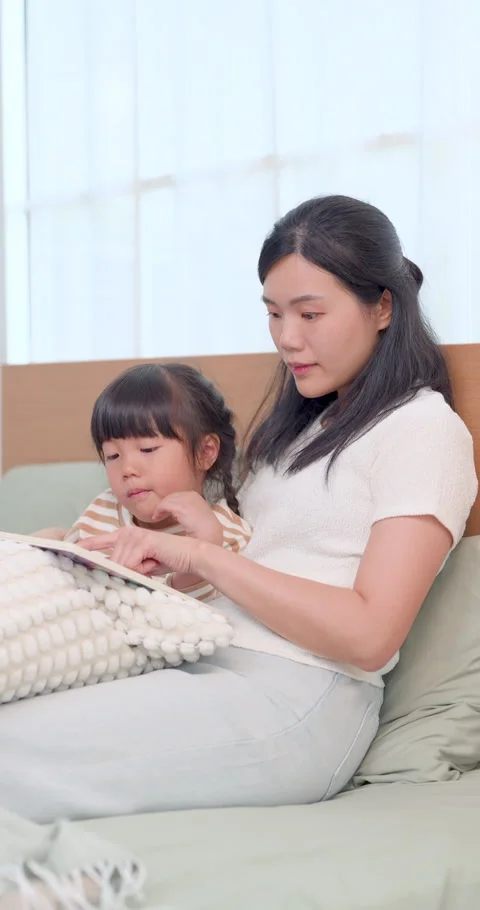 Asian mom points at page while preschool daughter listens on bed, creating Video stock 340967937
