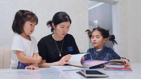 Asian mother reading book to two daughters at home, shared learning moment Stock Footage 333481640
