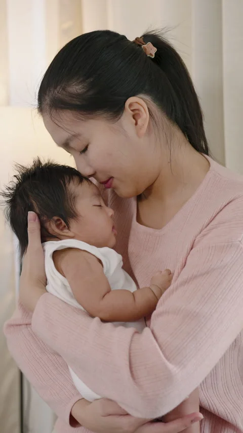 Asian mother smiling gently while kissing forehead of newborn baby boy lying in Stock Footage 326633687
