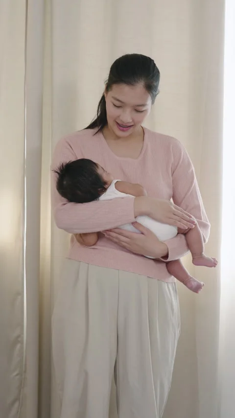 Asian mother smiling while holding baby boy gently on lap, newborn resting in Stock-Footage 333659988