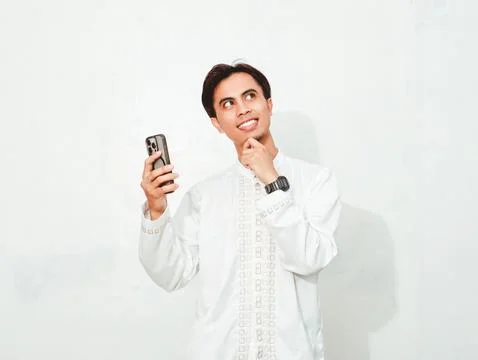 Asian Muslim man smiling while holding smartphone and looking up with think.. Stock Photos