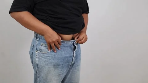 Asian Plus-size man buttoning oversized pants, celebrating weight loss. dieting Stock Footage 342475747