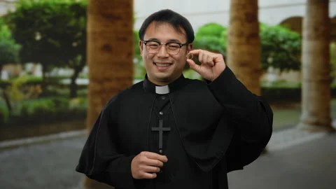 Asian priest holding a ring with a joyful smile stands outdoors in a church.. Vídeos de archivo 312006366