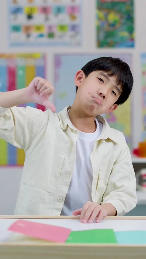 Asian schoolboy giving thumbs down in colorful classroom Stock Footage 326386008