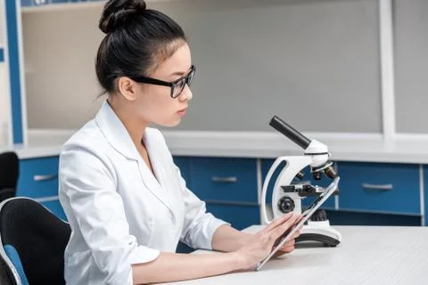 Asian scientist in lab coat with microscope and digital tablet working in che 库存照片