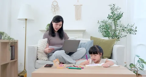 Asian single mother checking worksheet beside laptop while preschool daughter Stock Footage 340964171