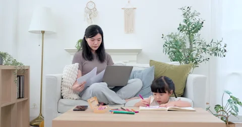 Asian single mother reviewing document on laptop while preschool daughter draws Stock Footage 332968869