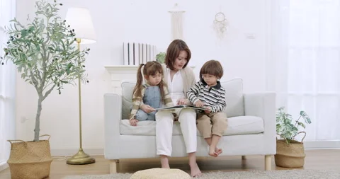 Asian single mother sits centered on white sofa, kids cuddle near, picture book Stock Footage 329546660