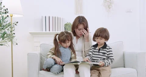 Asian single mother sits on sofa, turns picture book pages, guides toddler Stock Footage 339040613