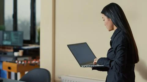 Asian software developer coming at desk and sitting down holding laptop with Stock Photos