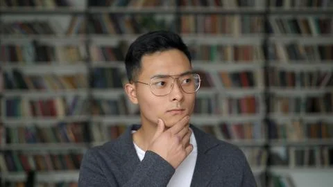 Asian student in grey jacket on background of blurred bookshelves in library 写真素材