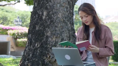 Asian Student Reading Book and Using Laptop Under Tree in Bangkok Park Stock Footage 334104804