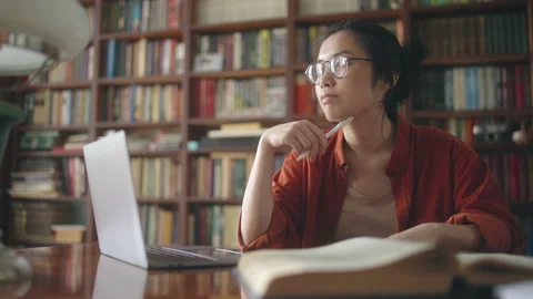 Asian student thinking over essay plot, working with books and pc at the library Vídeos de archivo 158930676