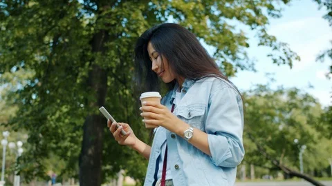 Asian student walking in park using smart phone drinking to go coffee smiling Stock Footage 116969466
