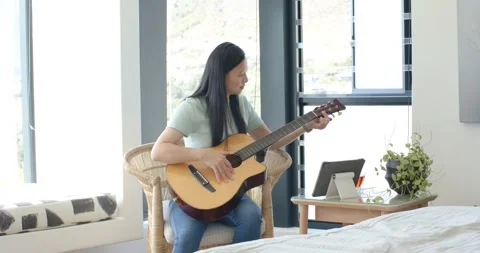 Asian woman consulting tablet tutorial picking up and tuning acoustic guitar in Stock Footage 337224921