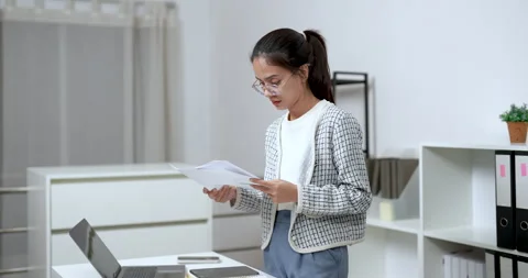 Asian woman reading documents while standing at desk during business task in 库存影片 336377441