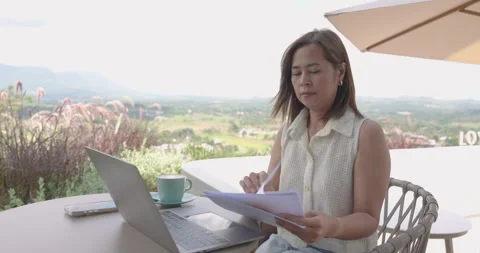 Asian woman reviewing document while sitting outdoor at cafe using laptop for Stock Footage 336378180