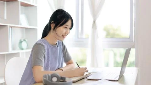 Asian woman taking notes in notebook while studying online in laptop at home, Stock Photos