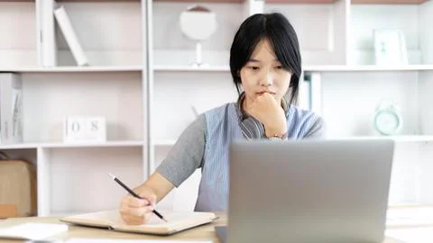Asian woman taking notes in notebook while studying online in laptop at home, 스톡 사진