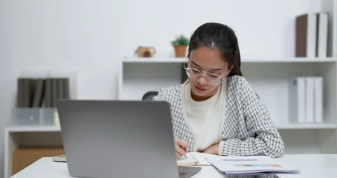 Asian woman taking notes while checking documents with laptop on white desk in Stock Footage 333602857