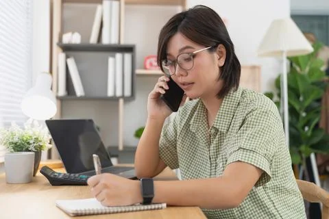 Asian woman taking notes while talking on smartphone at wooden desk with la.. Stock Photos