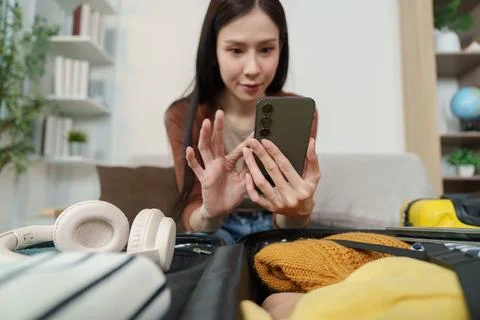 Asian woman using smartphone while packing suitcase for travel trip at home Stock Photos