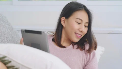 Asian woman using tablet drinking coffee while lying on the sofa at home. Stock Footage 110350457