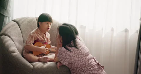 Asian young boy with Down syndrome playing ukulele guitar with mother on cozy ho Stock Footage 310175189