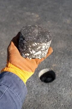 Asphalt core sample held in a gloved hand on a road surface. Hole from dril.. Stock Photos