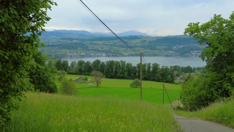Asphalt path leading down towards a green meadow with a lake and mountains in Stock Footage 334870410
