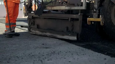 An asphalt paver throws fresh warm asphalt mixture close-up. Nearby a worker Stock Footage 261982949