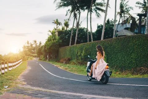 On the asphalt road of Bali on a motorcycle eating girl in a long white dress Stock Photos