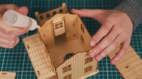 Assembling wooden house model with hands positioning roof section, applying glue 库存影片 335144133